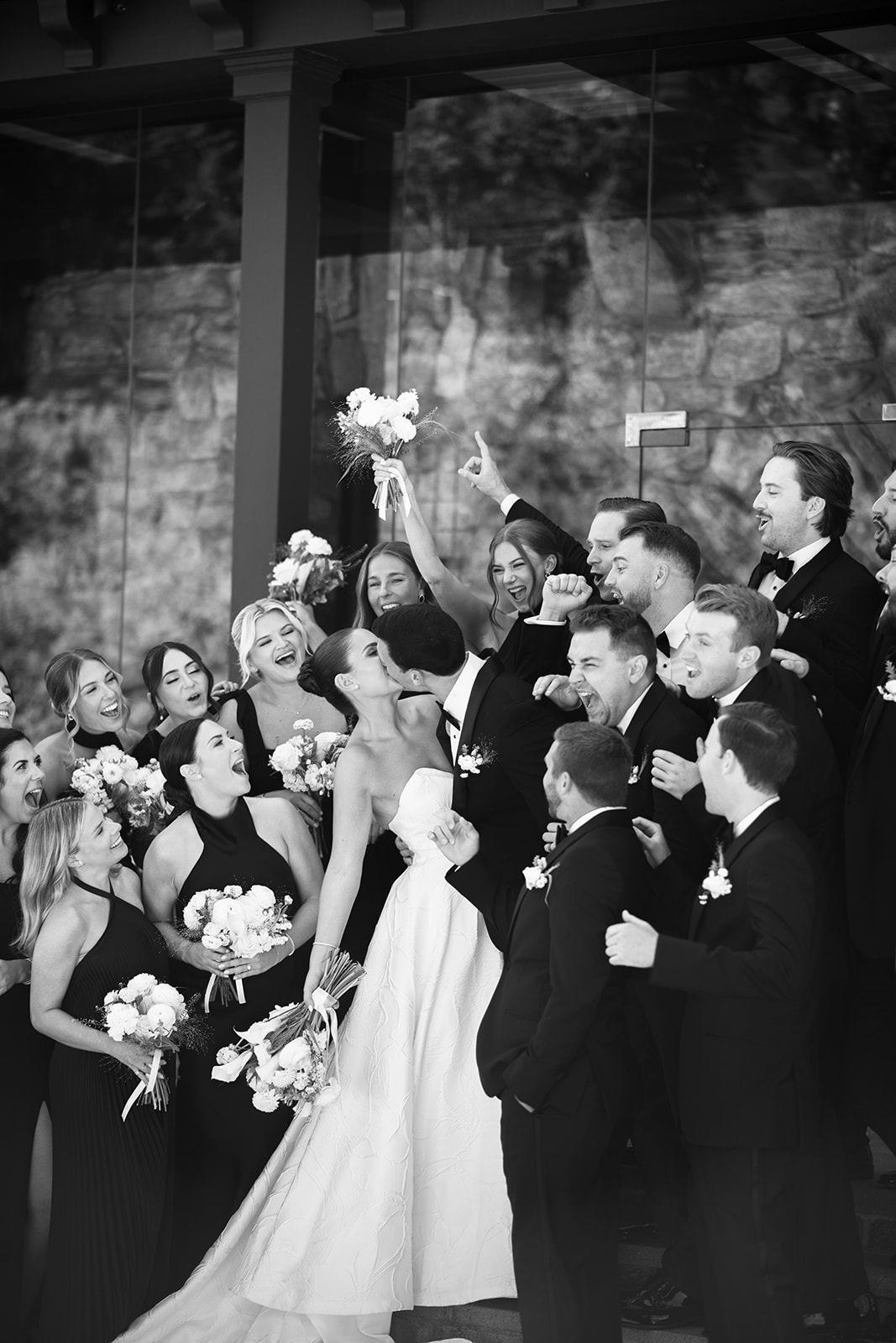 Black and white film photo of the bride and groom kissing surrounded by their cheering wedding party.